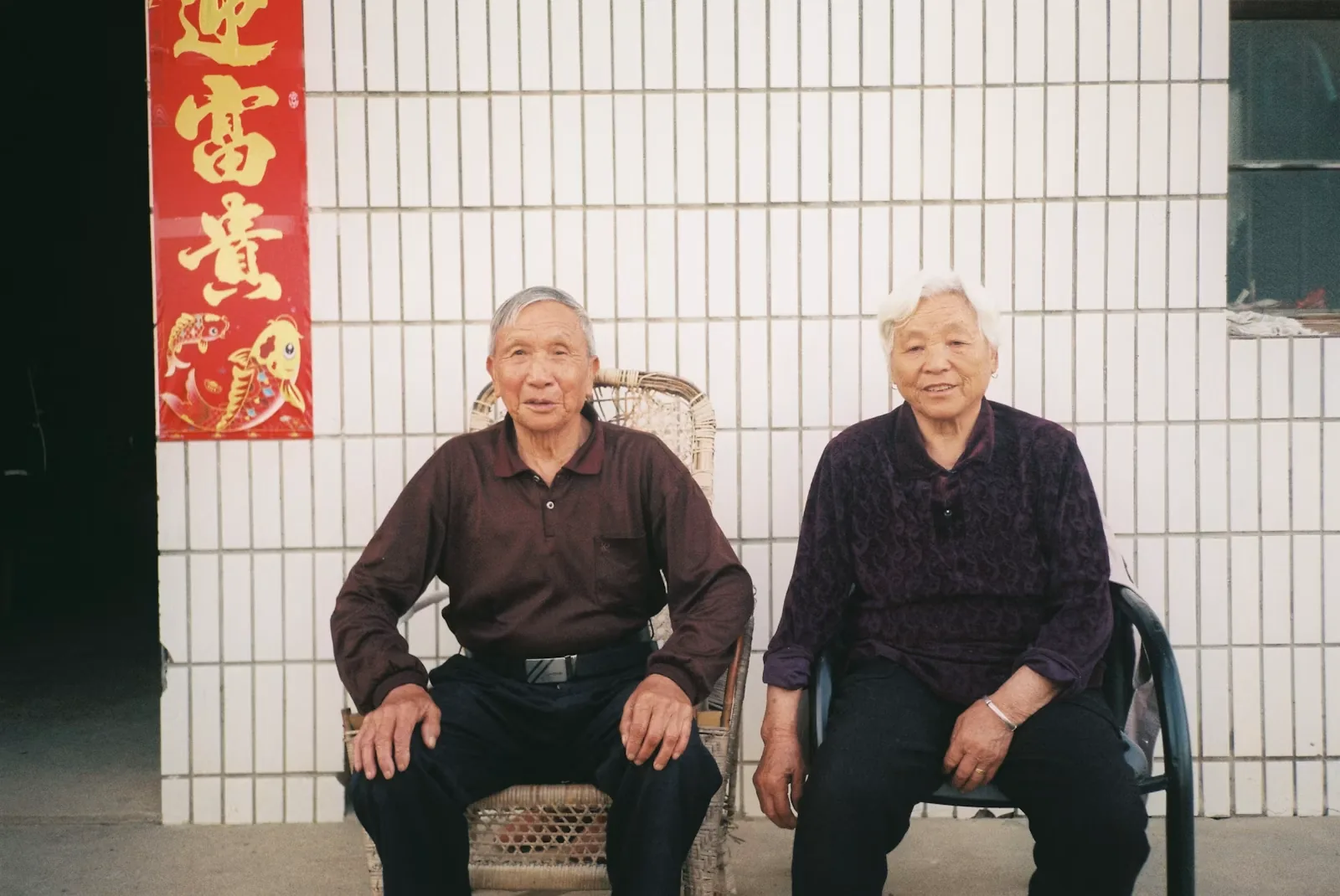 Elderly couple seated together in front of a white tiled wall