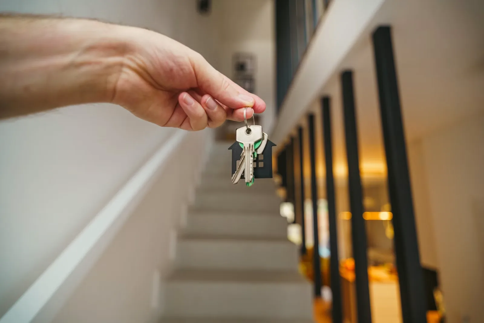 Hand holding house keys with a house-shaped keychain in front of a staircase